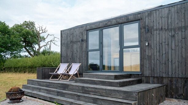 A modern building with a wooden facade, large glass windows, and a set of outdoor steps leading to a seating area with two lounge chairs. - Cosy Cabin at The Coppleridge 2 (Motcombe)