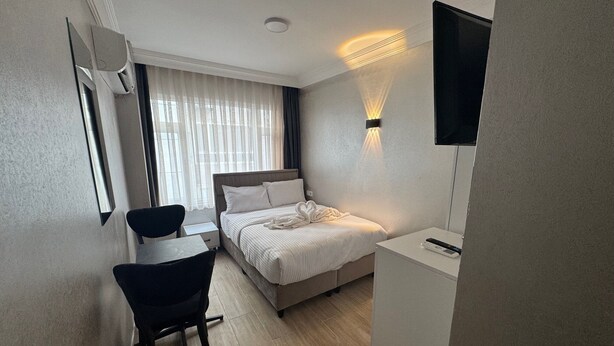 A hotel room with a bed, a desk with a chair, a TV mounted on the wall, and a window with curtains. - Old City Kadıköy Otel (Istanbul)