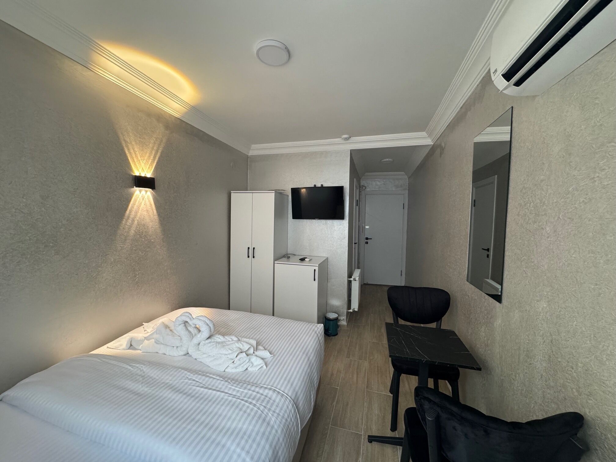 A hotel room with a bed, a desk, a chair, a wardrobe, a television, and an air conditioning unit.