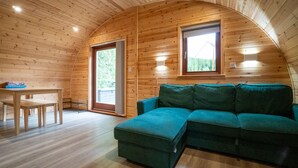 L’intérieur chaleureux d’un chalet avec des murs en bois, un canapé d’angle vert, une table en bois avec des chaises et deux fenêtres qui laissent entrer la lumière naturelle.