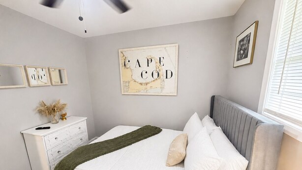 Um quarto com cama, cômoda, ventilador no teto, moldura com frase na parede e janela com persianas.