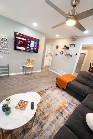 Un salon avec un canapé gris, une table basse, un téléviseur fixé au mur et un ventilateur de plafond.