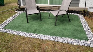 Uma área externa com espaço para sentar, com duas cadeiras e uma mesa, cercada por gramado e uma borda de pedrinhas, em frente a uma casa com janela e luzes de Natal.
