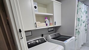 Uma lavanderia com duas máquinas de lavar brancas, uma prateleira com produtos de limpeza e um aviso indicando a finalidade do espaço.