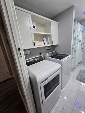 Een wasserette met twee witte wasmachines, een plank met schoonmaakmiddelen en een bordje met de functie van de ruimte.