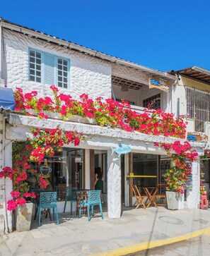 Un bâtiment à la façade blanche, aux volets bleus et orné de fleurs roses. Il y a des espaces de détente en plein air avec des chaises et des tables.