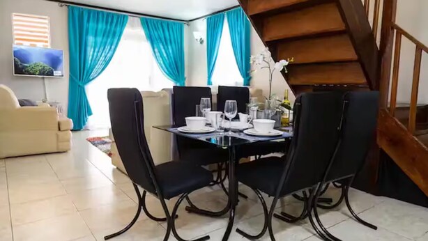 Un coin repas avec une table et des chaises noires, un téléviseur fixé au mur et des rideaux bleus.