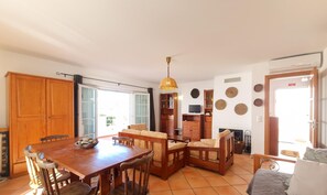 A spacious living area with a wooden dining table, chairs, a sofa, and a fireplace. There is a TV mounted on the wall and a ceiling fan. - Casa da Cestaria T2 Duplex by YHA (Cabanas de Tavira)