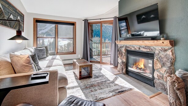 A cozy living room with a stone fireplace, a large TV, and a sofa with a coffee table. - Luxury Renovated 2BR | Walk to Slopes I River View (Keystone)