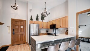 A modern kitchen with wooden cabinets, a stainless steel refrigerator, a central island with bar stools, and decorative trees. - Luxury Renovated 2BR | Walk to Slopes I River View (Keystone)