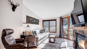 A modern living room with a leather armchair, a sofa, a fireplace, and a large window with a mountain view. - Luxury Renovated 2BR | Walk to Slopes I River View (Keystone)