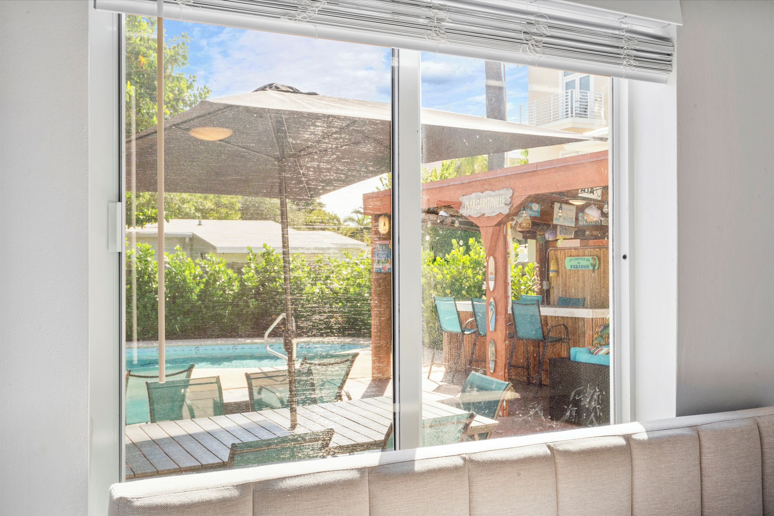 A view through a glass door of a pool area with a wooden deck, lounge chairs, and a bar area.