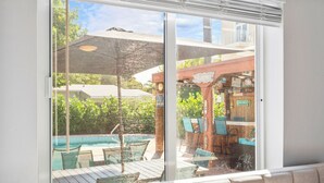 A view through a glass door of a pool area with a wooden deck, lounge chairs, and a bar area. - Beach Family Tiki Villa Walk to Beach Heated Pool (Pompano Beach)