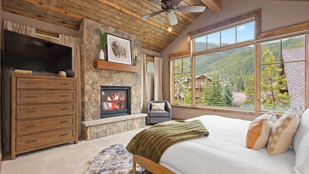 A bedroom with a stone fireplace, a large bed, a television, and a view of a mountainous landscape through large windows. - Alpine 28: Luxe Keystone Mountain Home - Sleeps 16 (Dillon)