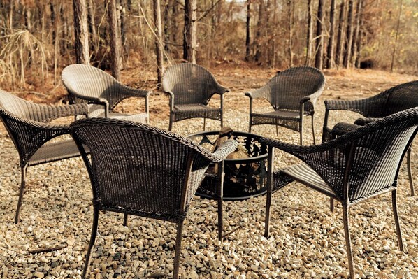 A circular outdoor seating area with wicker chairs and a central fire pit, surrounded by gravel and trees.