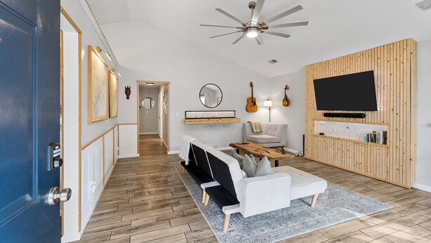 A modern living room with a sofa, coffee table, and TV. There is a wooden wall panel and a ceiling fan.