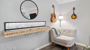 A modern living room with a marble-framed mirror, a wooden shelf with glasses, a guitar, a lamp, and a cushioned armchair.