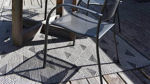 A wooden deck with a round table and metal chairs, a potted plant, and a yellow building in the background.
