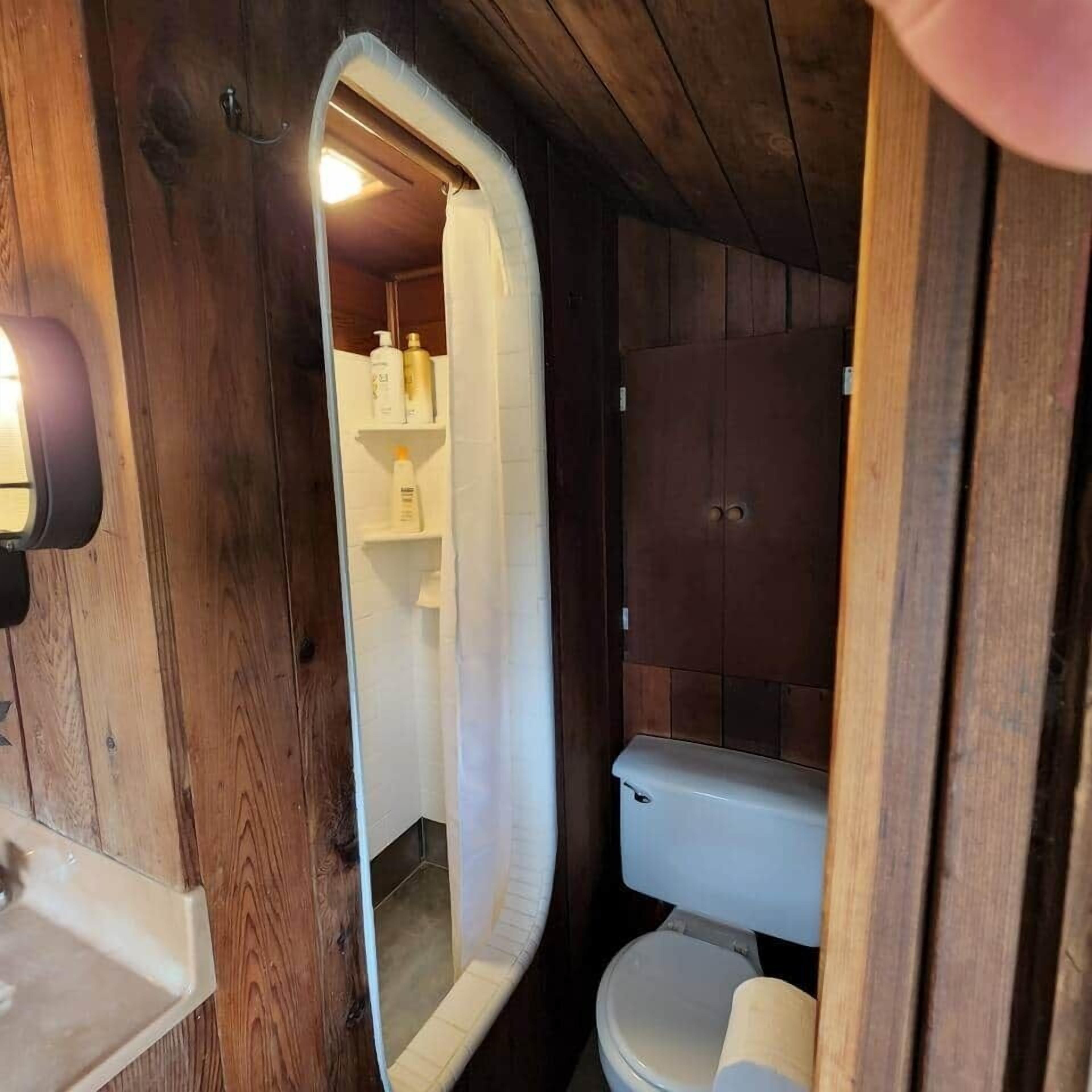Une petite salle de bain au décor intérieur en bois, avec des toilettes blanches et un miroir qui reflète l’espace de la douche.