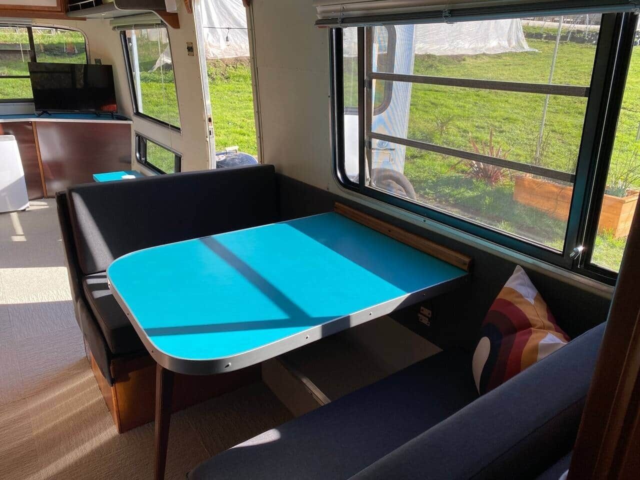 A compact RV interior with a blue table, a cushioned bench, and a window providing a view of the outdoors.