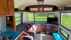 Interior compacto de autocaravana con kitchenette, sofá, una mesita y ventana con vistas a la vegetación.