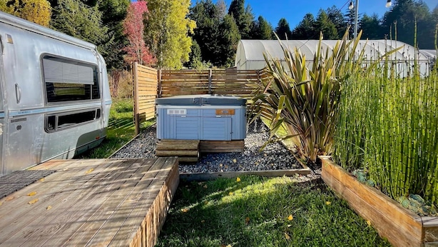 Un jardín con jacuzzi, terraza de madera y una caravana aparcada al lado.