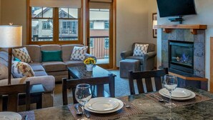 A cozy living room with a fireplace, a television mounted above it, and a dining area with a table set for two. - Relaxed Luxury 2BR at Grand Timber Lodge – Mountain-View Retreat in Breckenridge (Breckenridge)