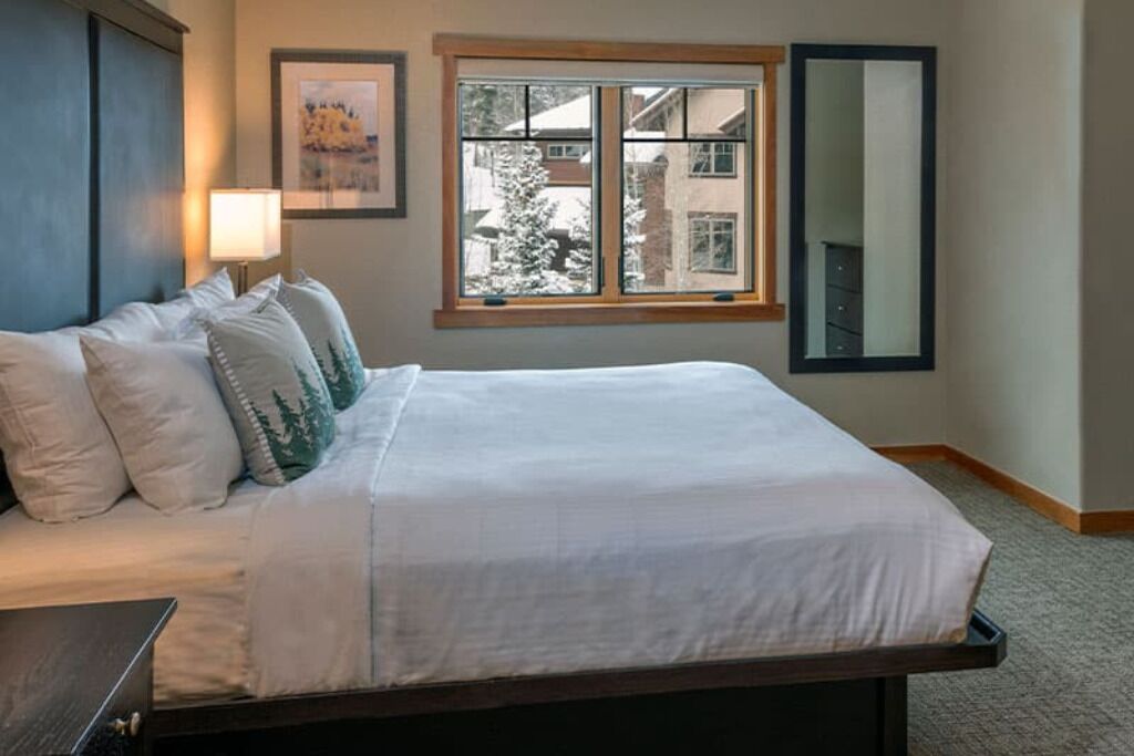 Un lit bien fait, avec du linge de lit blanc et des coussins décoratifs, une lampe de chevet, une fenêtre donnant sur des immeubles et un arbre, et un miroir accroché au mur.