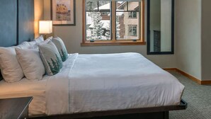 A neatly made bed with white linens and decorative pillows, a bedside lamp, a window with a view of buildings and a tree, and a mirror on the wall. - Relaxed Luxury 2BR at Grand Timber Lodge – Mountain-View Retreat in Breckenridge (Breckenridge)