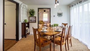 Comedor con una mesa de madera dispuesta para seis personas, rodeada de sillas con cojines estampados. Se ve una estantería y una planta en maceta al fondo.