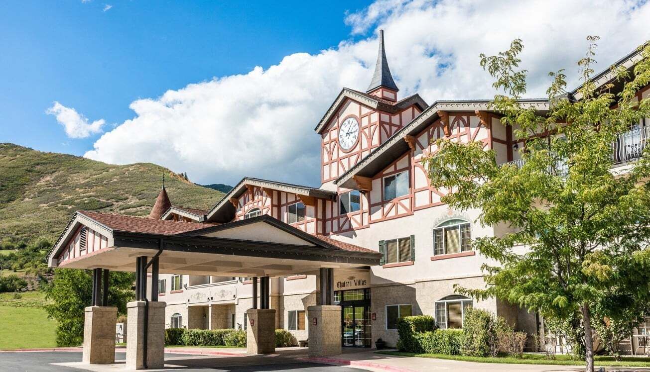 A traditional chalet-style hotel with a prominent clock tower, a covered entrance, and a mountainous backdrop.