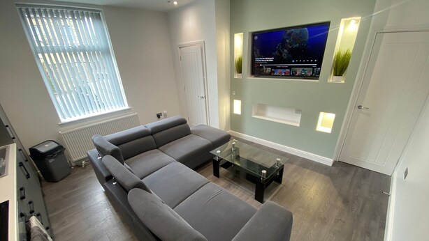 A modern living room with a grey sectional sofa, a glass coffee table, and a flat-screen TV mounted on the wall.