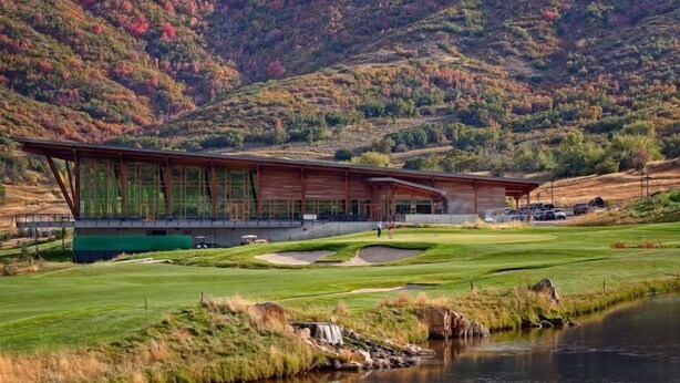 Un edificio moderno con una amplia fachada de vidrio, ubicado junto a un campo de golf y una masa de agua.