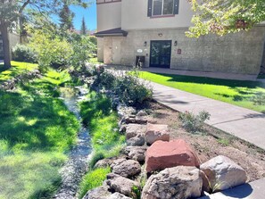 Un área ajardinada con un pequeño arroyo, rocas y un edificio de fachada de piedra y puerta.