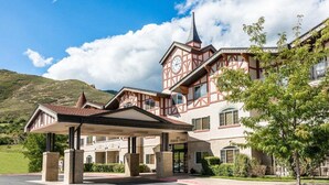 A traditional chalet-style hotel with a prominent clock tower, a covered entrance, and a mountainous backdrop.