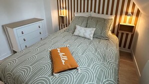 Un dormitorio con techo inclinado, decoración de pared a rayas, una cama con ropa de cama estampada, una mesita de noche con lámpara y un aparador pequeño.