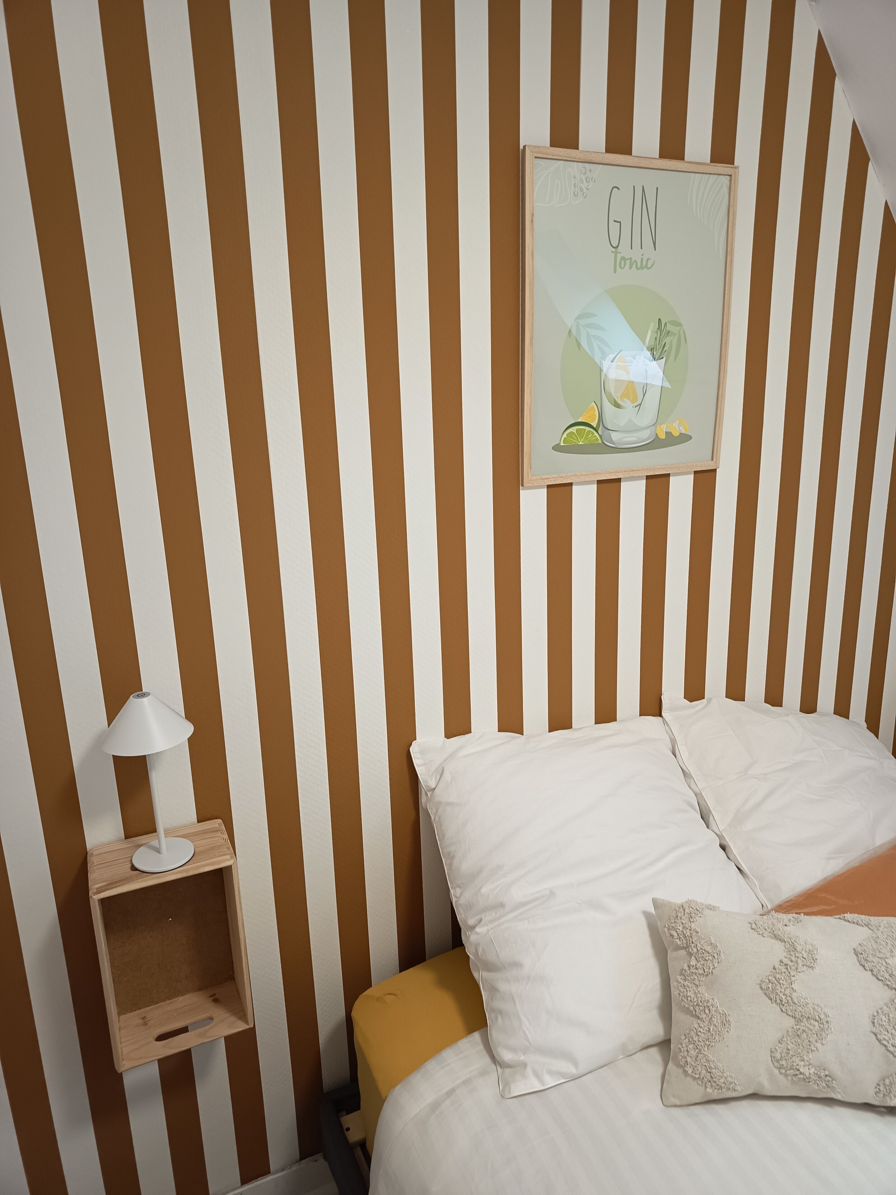 A striped wall with a framed picture of gin tonic, a bedside table with a lamp, and a bed with pillows.