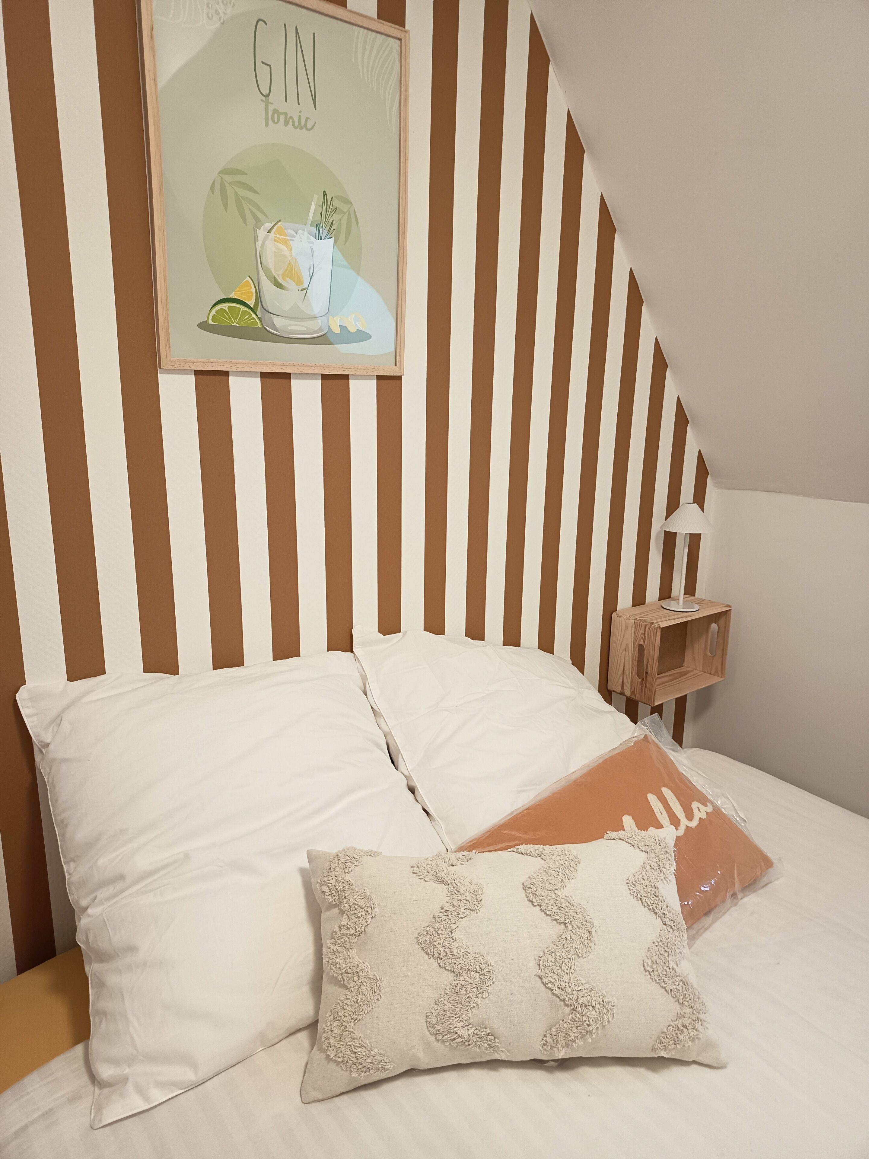 Ein Bett mit weißen und beigen Kissen, ein gerahmtes Bild an der Wand und ein Nachttisch mit einer Lampe.