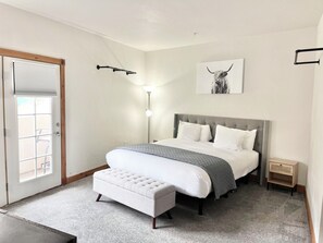 Une chambre à coucher comprenant un lit, des tables de chevet, un banc, une porte avec une fenêtre et un luminaire fixé au mur.