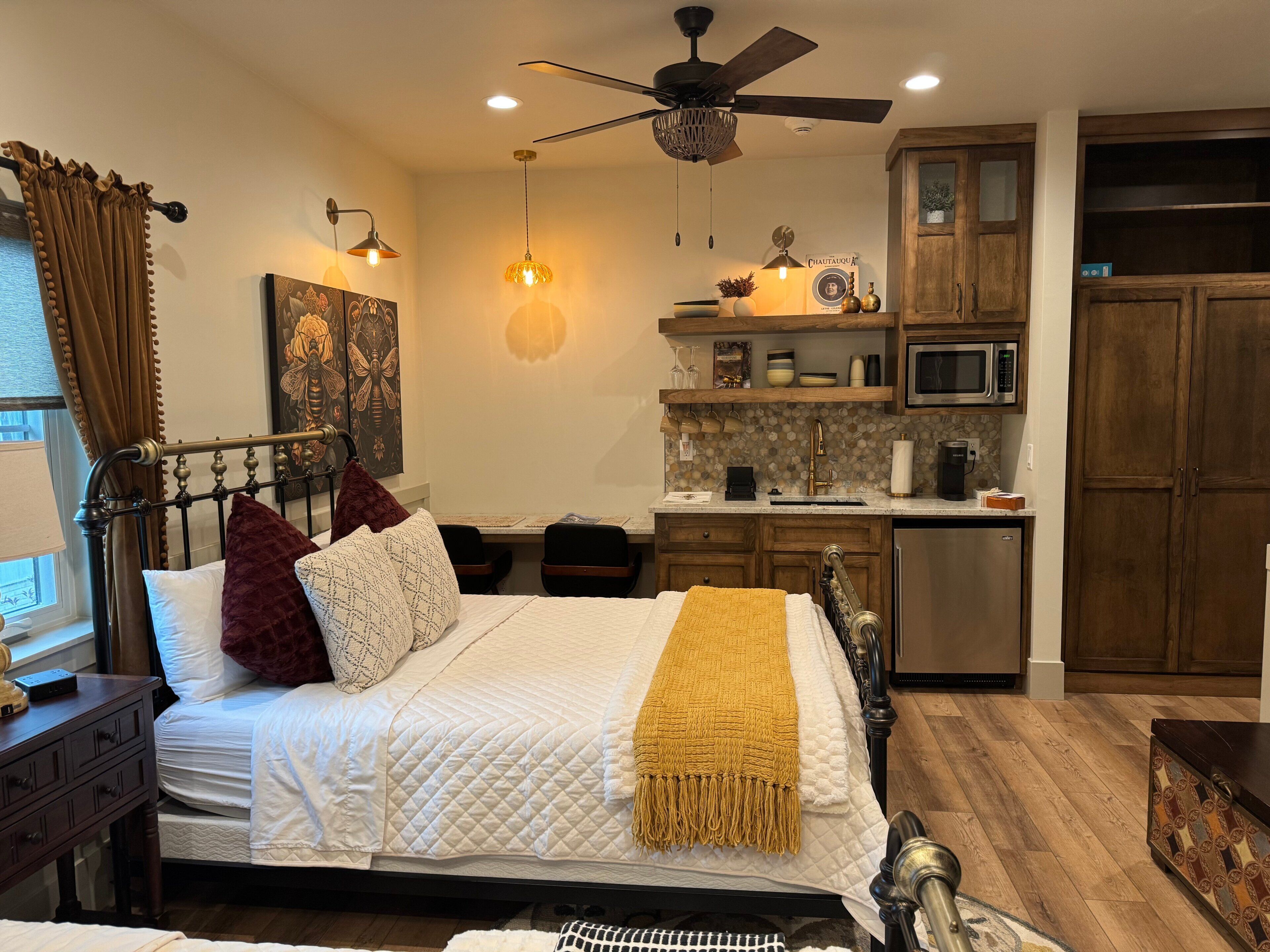 A bedroom with a bed, bedside tables, a desk, a chair, a ceiling fan, a kitchen area with a microwave, and cabinets.