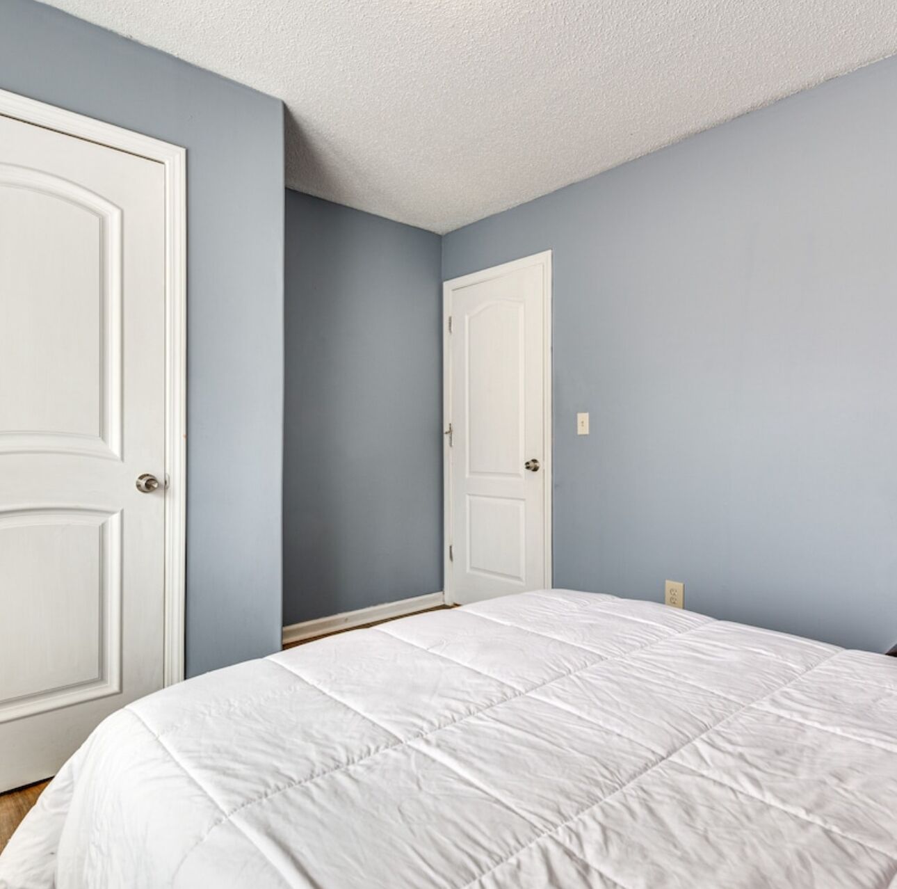 A bedroom with two white doors, a white bedspread, and light blue walls.