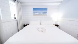 A hotel room with a large bed, white walls, a painting of a beach, and two bedside tables with lamps. - The Shoreline Studio - Perfect AC Getaway! (Atlantic City)