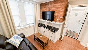 A compact living space with a brick wall, a flat-screen TV, a dining area with a black countertop, and a white door with a 'No Smoking' sign. - The Shoreline Studio - Perfect AC Getaway! (Atlantic City)