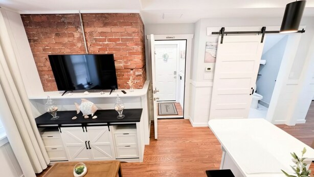 A modern living room with a brick wall, a flat-screen TV, white cabinets, and a sliding door leading to another room. - The Shoreline Studio - Perfect AC Getaway! (Atlantic City)