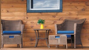 Two wicker chairs with blue cushions and a small table with a yellow vase between them, against a wooden wall with a window above.