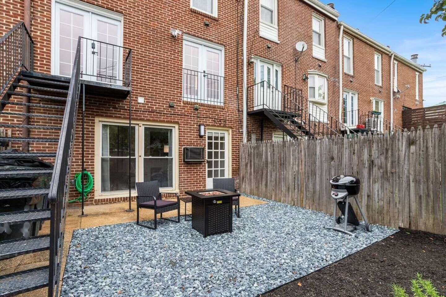 A brick apartment with a patio area featuring a grill, outdoor furniture, and a wooden fence.