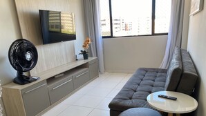 A modern living room with a grey sofa, a small round table, a black standing fan, and a TV mounted on the wall. - 02 QUARTOS próximo a PRAIA de PAJUÇARA (Maceió)