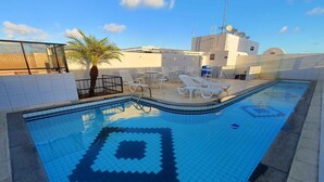 A swimming pool with a geometric tile pattern, surrounded by white lounge chairs and a palm tree. - 02 QUARTOS próximo a PRAIA de PAJUÇARA (Maceió)