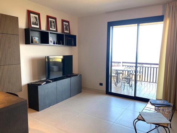 Una sala de estar moderna con un televisor de pantalla plana, una estantería con fotografías enmarcadas y un balcón con mobiliario exterior.