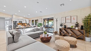 A modern living room with a grey sectional sofa, a wooden coffee table, and woven poufs. A kitchen is visible in the background with a dining area and large windows. - Large Private Backyard Oasis. Family Friendly - Arcade Room-5min to Down Town PS (Palm Springs)